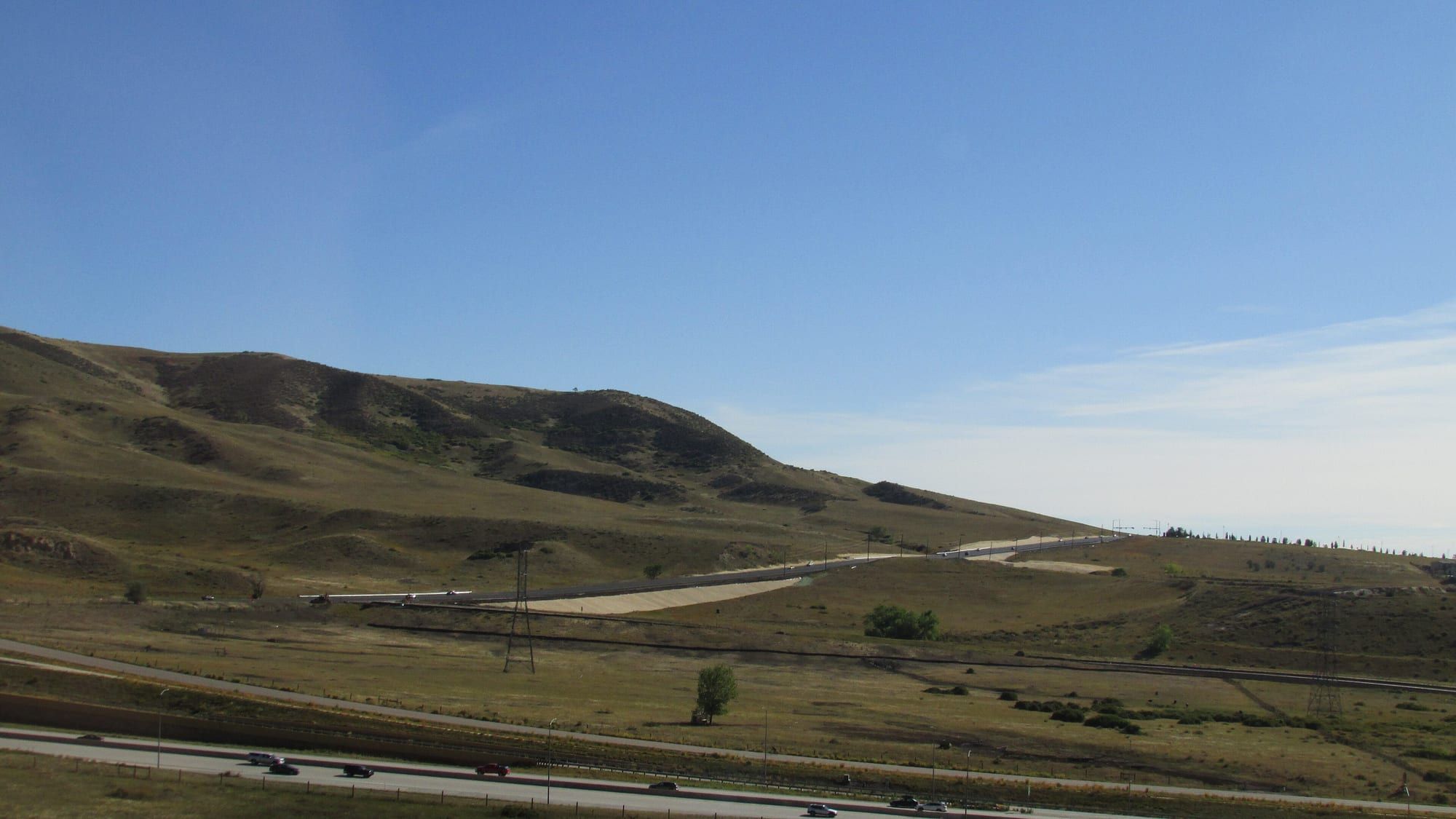 Brown and green grassy hills under a clear blue sky, with a highway and power lines running along their foot